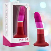 The Avant Blush Avant P3 Pride Silicone Dildo with Suction Cup, featuring pink, red, and white stripes, stands beside its "PRIDE by AVANT" packaging as colorful lights blur in the background.